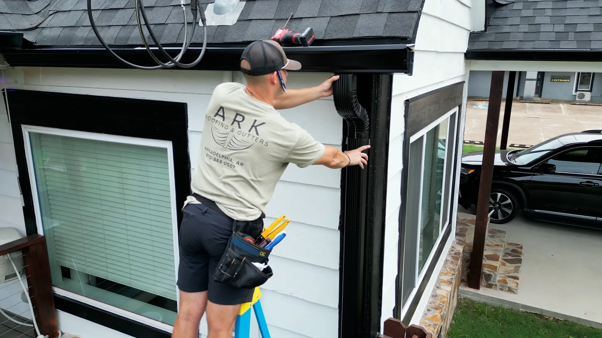 Ark Roofing crew member fitting a black downspout beside a picture window